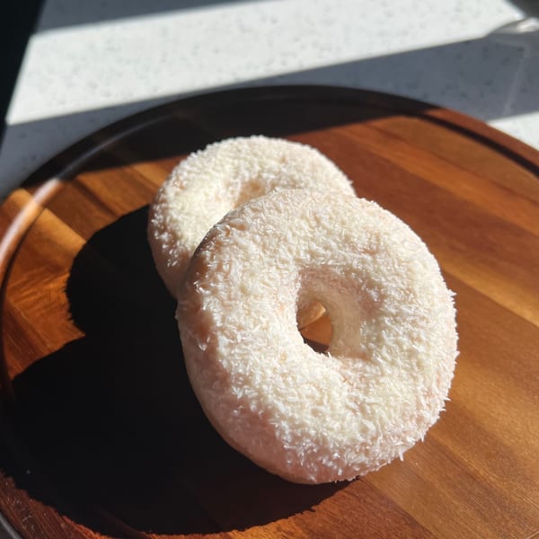 Two coconut-flaked donuts on a wooden tray, bathed in sunlight and shadow.
